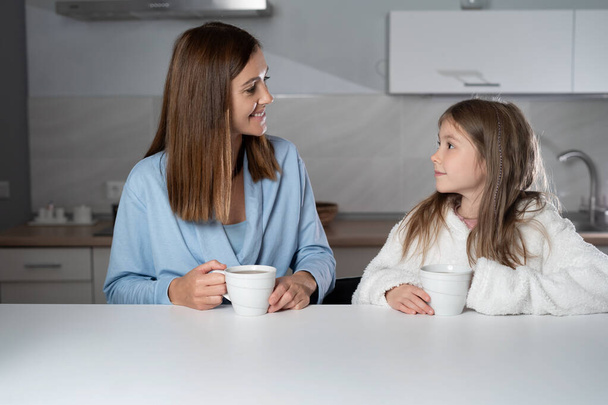 Im Rahmen sitzt eine gut gelaunte Mutter mit ihrer Tochter am Küchentisch und trinkt morgens heißen Tee. Neugieriges Mädchen fragt Mama etwas - Foto, Bild