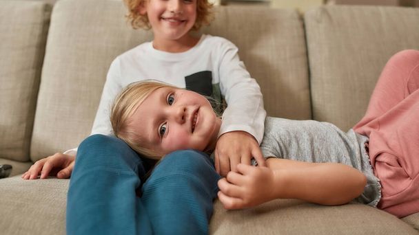 Portrait of cute little girl smiling at camera while spending time together with her sibling brother, cuddling on a sofa at home - Photo, image