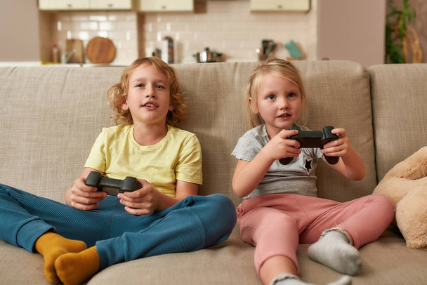Leisure time. Cheerful kids, little boy and girl playing video games using joystick or controller while sitting together on sofa at home - Fotó, kép