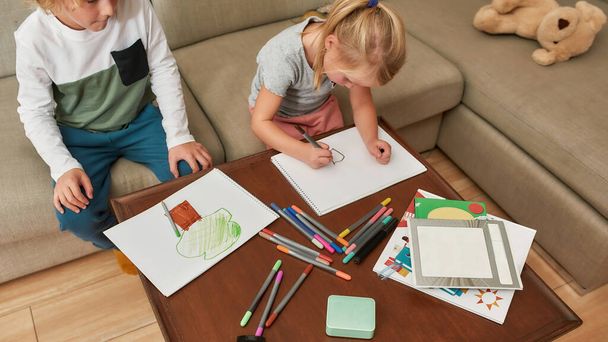 Cropped shot of little kids, boy and girl drawing on paper using marker pen, sitting together on a sofa at home - 写真・画像