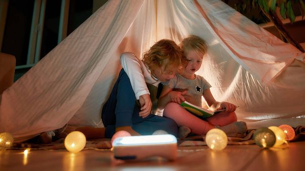Duas crianças pequenas, irmão e irmã lendo um livro juntos enquanto sentados em um cobertor em uma tenda feita com lençóis em casa - Foto, Imagem