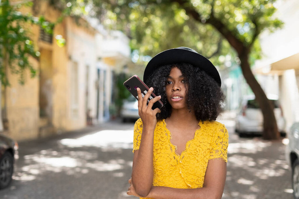 young black woman with curly hair, with red mask, yellow dress, using her smart phone - Foto, Imagen
