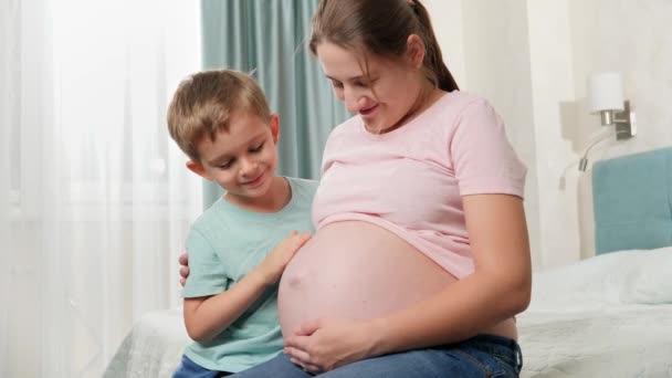 妊娠中の母親の大きな腹に触れるかわいい男の子がベッドルームに座っています。赤ちゃんを期待している家族の肯定的な感情 - 映像、動画