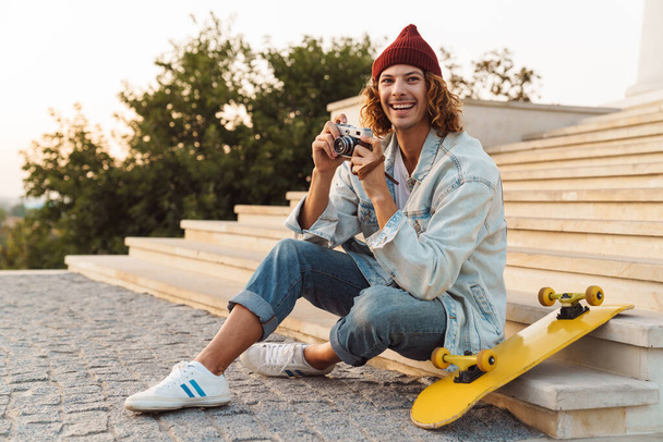 Picture of positive happy young curly man scater sitting outdoors and holding camera - Photo, image
