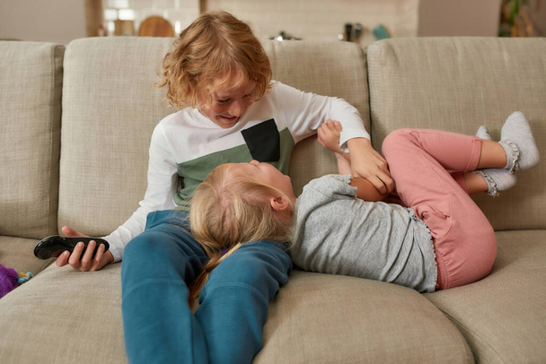 Lovely little siblings, boy and girl having fun, playing together while watching TV, cuddling on a sofa at home - Foto, Bild