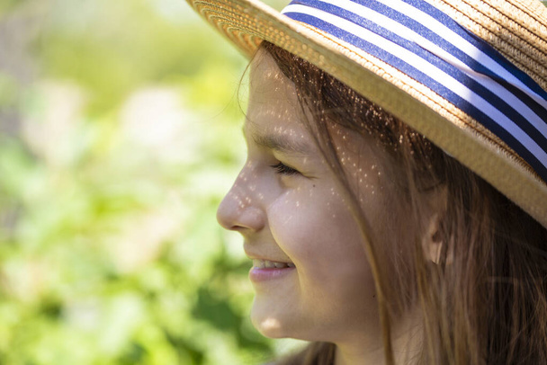 Uśmiechnięta dziewczyna z długimi włosami w słomkowym kapeluszu z bliska na tle letniego ogrodu. - Zdjęcie, obraz