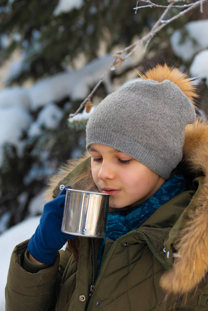 Une fille dans un chapeau d'hiver et une veste avec de la fourrure boit le thé chaud d'une tasse en métal en hiver dans la forêt. Le concept des promenades hivernales. - Photo, image