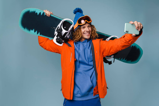 Przystojny szczęśliwy sportowiec robi selfie na komórce odizolowany na niebieskim tle, w zimowej kurtce, trzymając snowboard - Zdjęcie, obraz