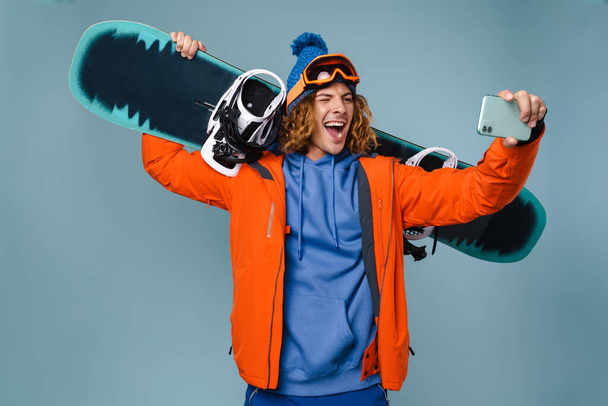 Handsome happy sportsman taking selfie on cellphone isolated over blue background, wearing winter jacket, holding snowboard - Fotó, kép
