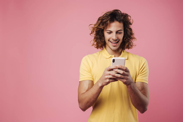 Portrait of an excited young man using mobile phone isolated over pink background - Foto, Imagem