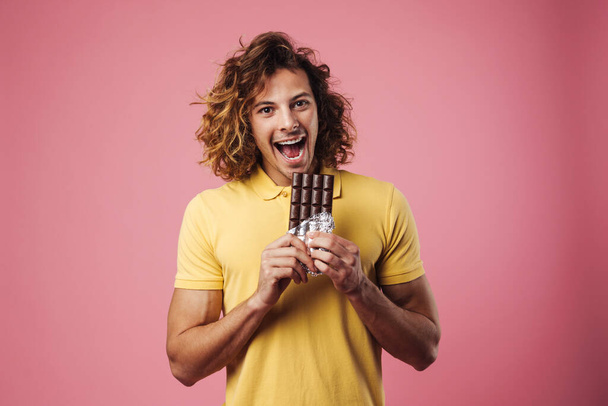 Cheerful handsome guy smiling while eating chocolate isolated over pink background - Фото, изображение