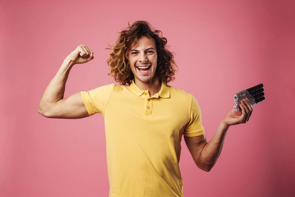 Cheerful handsome guy showing his bicep while holding chocolate isolated over pink background - Fotó, kép