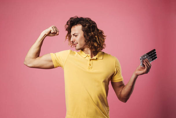 Confident healthy young man flexing biceps while showing chocolate bar isolated over pink background - 写真・画像