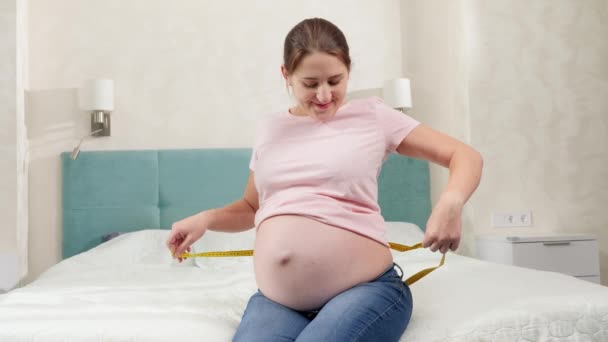 Heureuse femme enceinte souriante assise sur le lit et mesurant son gros abdomen avec du ruban à mesurer - Séquence, vidéo