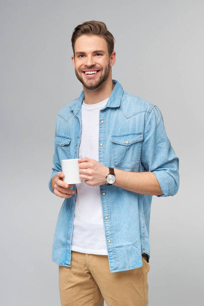 Portrait of young handsome caucasian man in jeans shirt over light background holding cup of coffee - Foto, Imagem