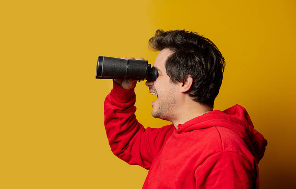 Smiling white guy in rd hoodie with binocular on yellow background - Foto, immagini