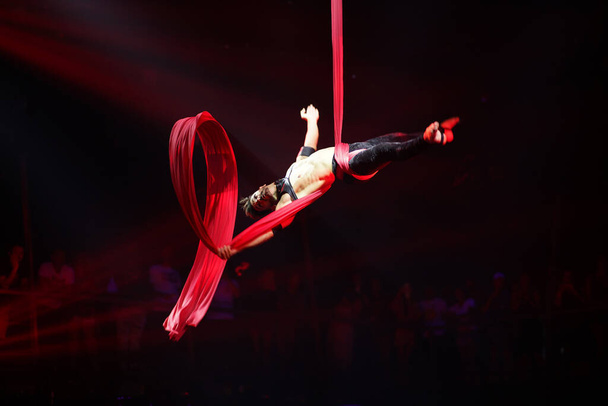 Brazilský silný sexy muž vystupuje na leteckém hedvábí v červených světlech, kapky na leteckém hedvábí. Sportovní trénink tělocvična a životní styl koncept. - Fotografie, Obrázek