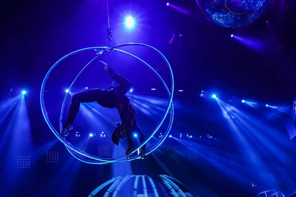 Flexible young woman make performance on aerial hoop, flexible back on aerial hoop, aerial circus show, blue light. Flexible woman gymnast upside down on hoop. Night club performance - Photo, Image