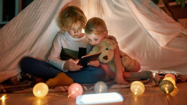 Crianças adoráveis, menino e menina usando tablet digital enquanto passam o tempo juntos, sentados em uma cabana feita com lençóis em casa - Foto, Imagem