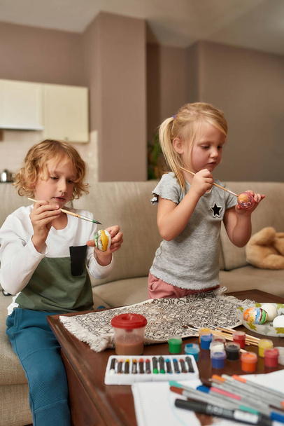 Portrait of two cute little children, brother and sister looking focused while painting colorful Easter eggs, sitting on a couch at home - Fotografie, Obrázek