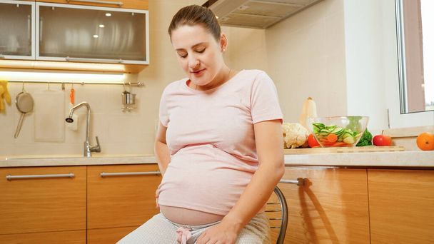 Fiatal terhes nő szenved a hasi fájdalom ül és pihen a széken a konyhában. Fájdalom és fájdalom a terhesség és a szülés alatt - Fotó, kép