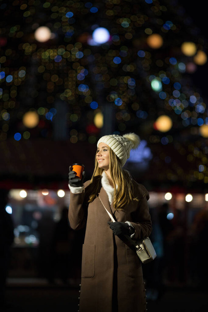 beautiful girl, in the evening, against the background of lights with a drink in an orange glass, a white hat and a beige coat. smiling at camera, close-up portrait - Foto, Bild