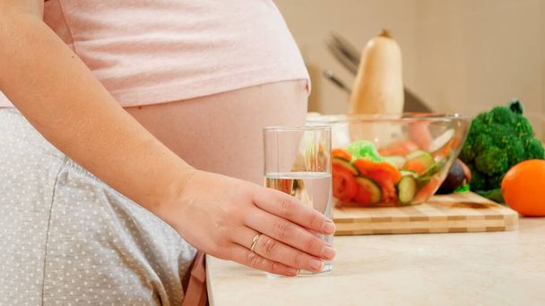 Closeup of pregnant woman expecting baby taking glass of water from kitchen tabletop. Concept of healthy lifestyle, nutrition and hydration during pregnancy - Fotoğraf, Görsel