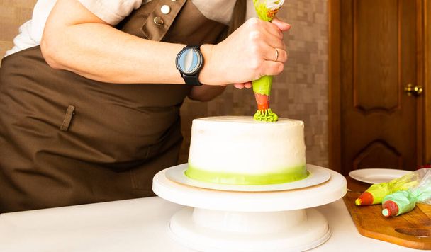 Woman using confectioners bag for putting whipped cream of green color and decorating Birthday cake at home. Concept of homemade cooking and decorating cake. Female confectioner making cake. - 写真・画像
