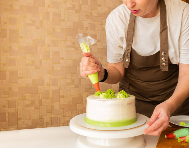 Woman using confectioners bag for putting whipped cream of green color and decorating Birthday cake at home. Concept of homemade cooking and decorating cake. Female confectioner making cake. - Fotografie, Obrázek
