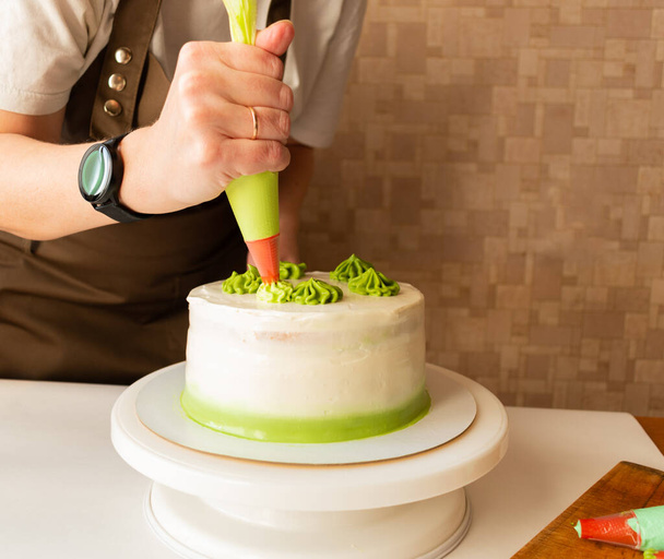 Woman using confectioners bag for putting whipped cream of green color and decorating Birthday cake at home. Concept of homemade cooking and decorating cake. Female confectioner making cake. - Фото, изображение