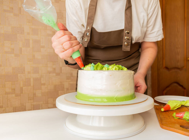 Woman using confectioners bag for putting whipped cream of green color and decorating Birthday cake at home. Concept of homemade cooking and decorating cake. Female confectioner making cake. - Φωτογραφία, εικόνα