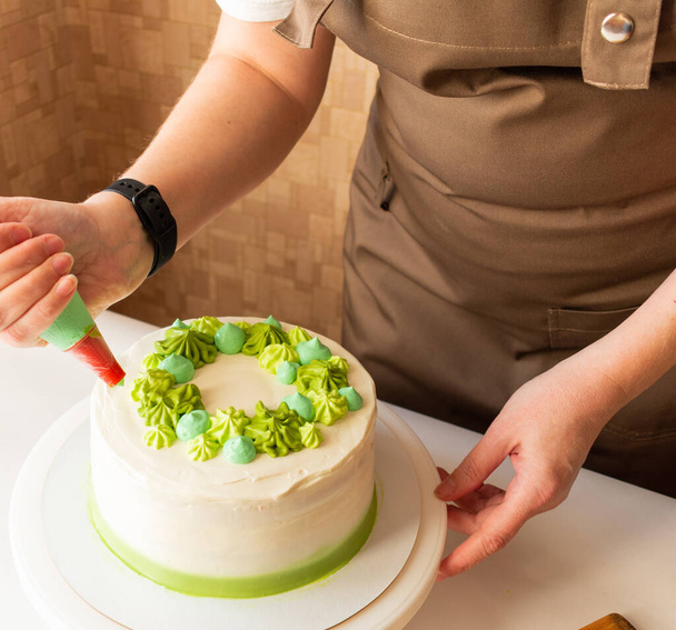 Woman using confectioners bag for putting whipped cream of green color and decorating Birthday cake at home. Concept of homemade cooking and decorating cake. Female confectioner making cake. - Zdjęcie, obraz