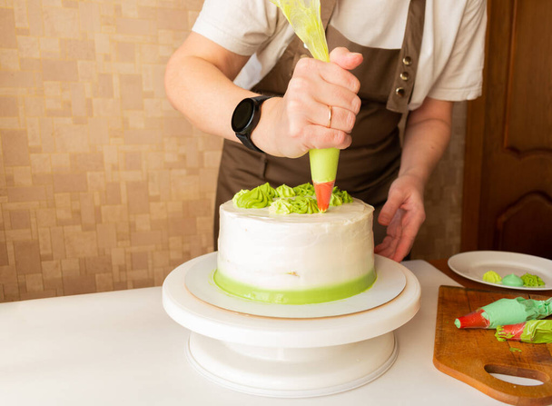 Woman using confectioners bag for putting whipped cream of green color and decorating Birthday cake at home. Concept of homemade cooking and decorating cake. Female confectioner making cake. - Valokuva, kuva
