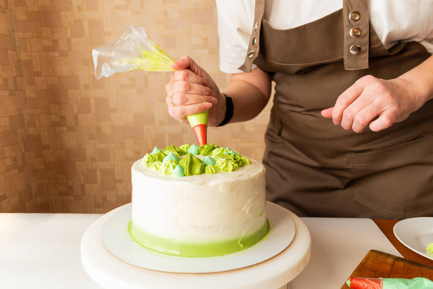 Woman using confectioners bag for putting whipped cream of green color and decorating Birthday cake at home. Concept of homemade cooking and decorating cake. Female confectioner making cake. - Фото, изображение