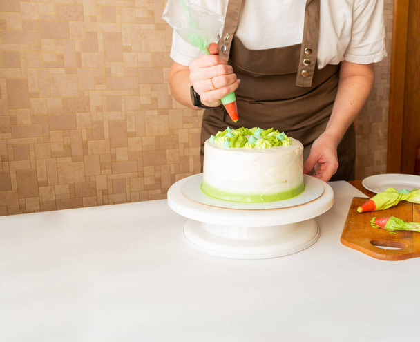 Woman using confectioners bag for putting whipped cream of green color and decorating Birthday cake at home. Concept of homemade cooking and decorating cake. Female confectioner making cake. - Foto, Bild