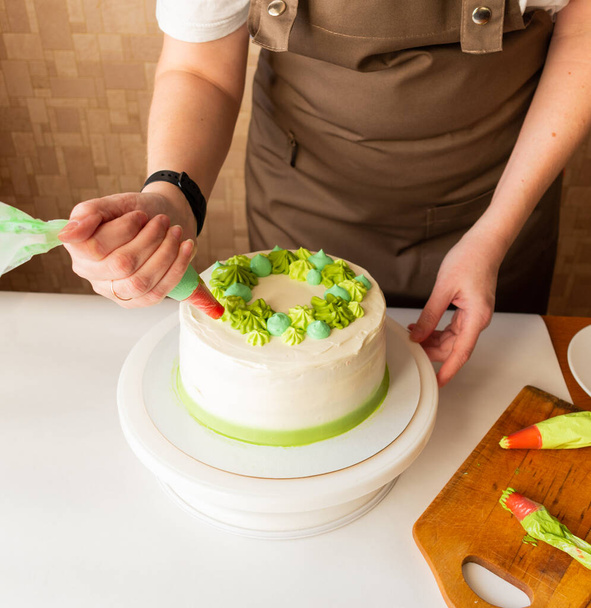 Woman using confectioners bag for putting whipped cream of green color and decorating Birthday cake at home. Concept of homemade cooking and decorating cake. Female confectioner making cake. - Фото, зображення