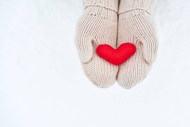 Holding red little soft fabric toy heart in beige mittens against background of white freshly fallen snow. Creative greeting card for Valentines Day. Empty space for your greeting text. - Photo, Image