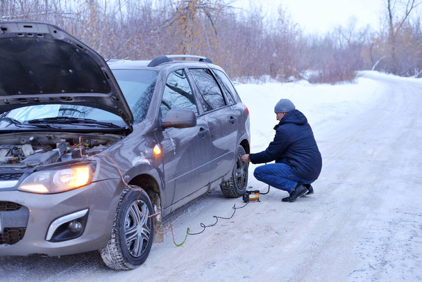 Hava basıncını kontrol eden ve lastiklerdeki havayı otomobil eletric pompasıyla dolduran bir adam kışın ormanda karlı bir yolda pompa kullanıyor. Bozuk araba konsepti. - Fotoğraf, Görsel