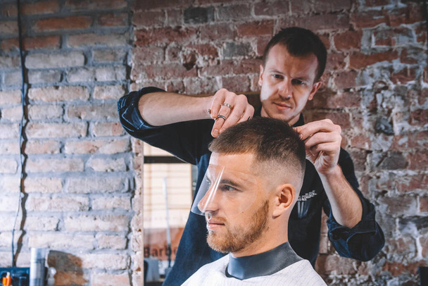 Młody fryzjer goli głowę klienta elektrycznym przycinaczem. Młody brodaty fryzjer w salonie fryzjerskim. Koncepcja sklepu reklamowego i fryzjerskiego. - Zdjęcie, obraz