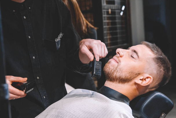 Mladý holič holí klientovi vousy elektrickým střihačem. Mladý vousatý muž se nechal ostříhat kadeřníkem v holičství. Koncept reklam a holičství. - Fotografie, Obrázek