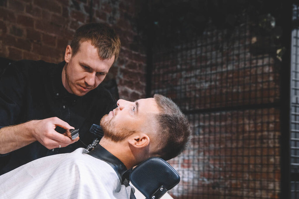 Joven peluquero macho afeita la cabeza del cliente con un trimmer eléctrico. Joven barbudo cortado por peluquería en la peluquería. Concepto de publicidad y peluquería. - Foto, imagen
