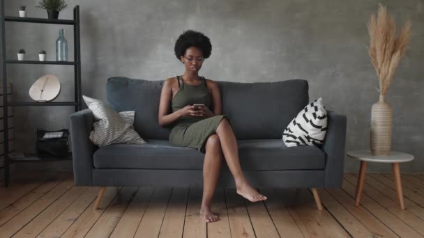 Gelukkig Afrikaans Amerikaanse jonge vrouw zitten ontspannen op gezellige bank blij om te verhuizen naar een nieuw appartement. Glimlachend zwart meisje rust op comfortabele bank in de woonkamer dromen - Video