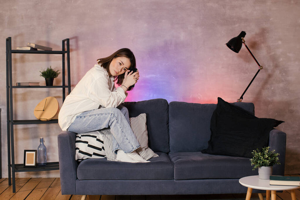 Junge asiatische Frau sitzt auf einem Sofa in einem gemütlichen Zimmer und schaut in die Kamera - Foto, Bild