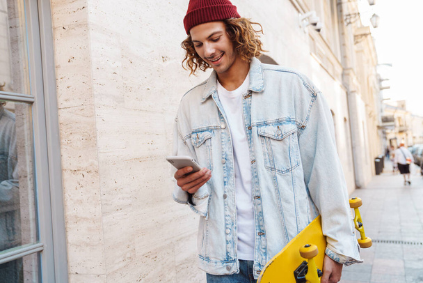Photo of a happy young curly man walking outdoors by street with skateboard and using mobile phone - Photo, Image