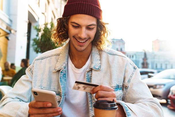 Image of cheery positive young curly man sitting in cafe outdoors and using mobile phone while holding credit card - Valokuva, kuva