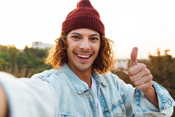 Image of a happy cheerful young curly man walking outdoors by street and taking a selfie by camera and showing thumbs up - Valokuva, kuva