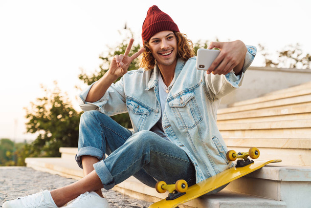 Image of cheery young curly man scater sitting outdoors and taking a selfie by mobile phone and showing peace gesture - Foto, Imagem