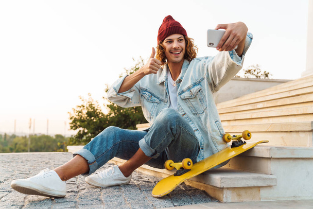Image of pleased young curly man scater sitting outdoors and taking a selfie by mobile phone while showing thumbs up gesture - Foto, Imagen