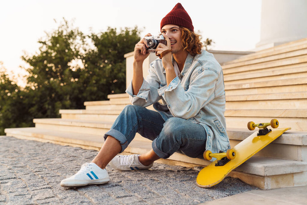Obrázek pozitivního veselého mladého kudrnatého muže, jak sedí venku a drží fotoaparát - Fotografie, Obrázek