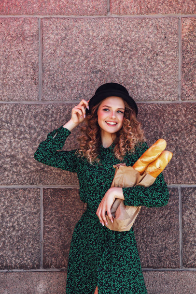 Portrait pretty young woman with baguette in hands on background texture of wall. Girl dressed in French style show emotion with bread. Female in stylish clothes holding fresh baguettes. Copy space - Foto, imagen